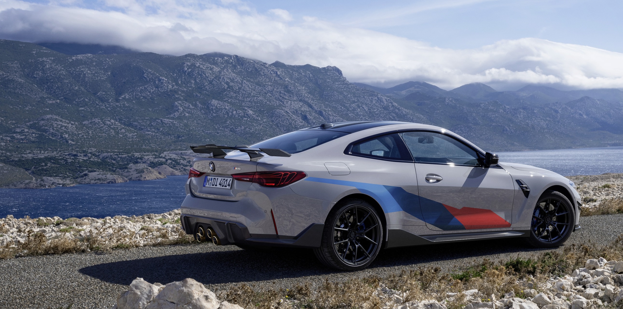A BMW M4 sits on a road overlooking a lake and mountain range.