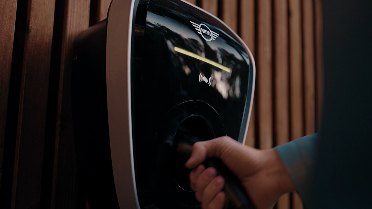 A MINI Wallbox charger is mounted to the wall. A hand is grabbing the handle of the charging cable.