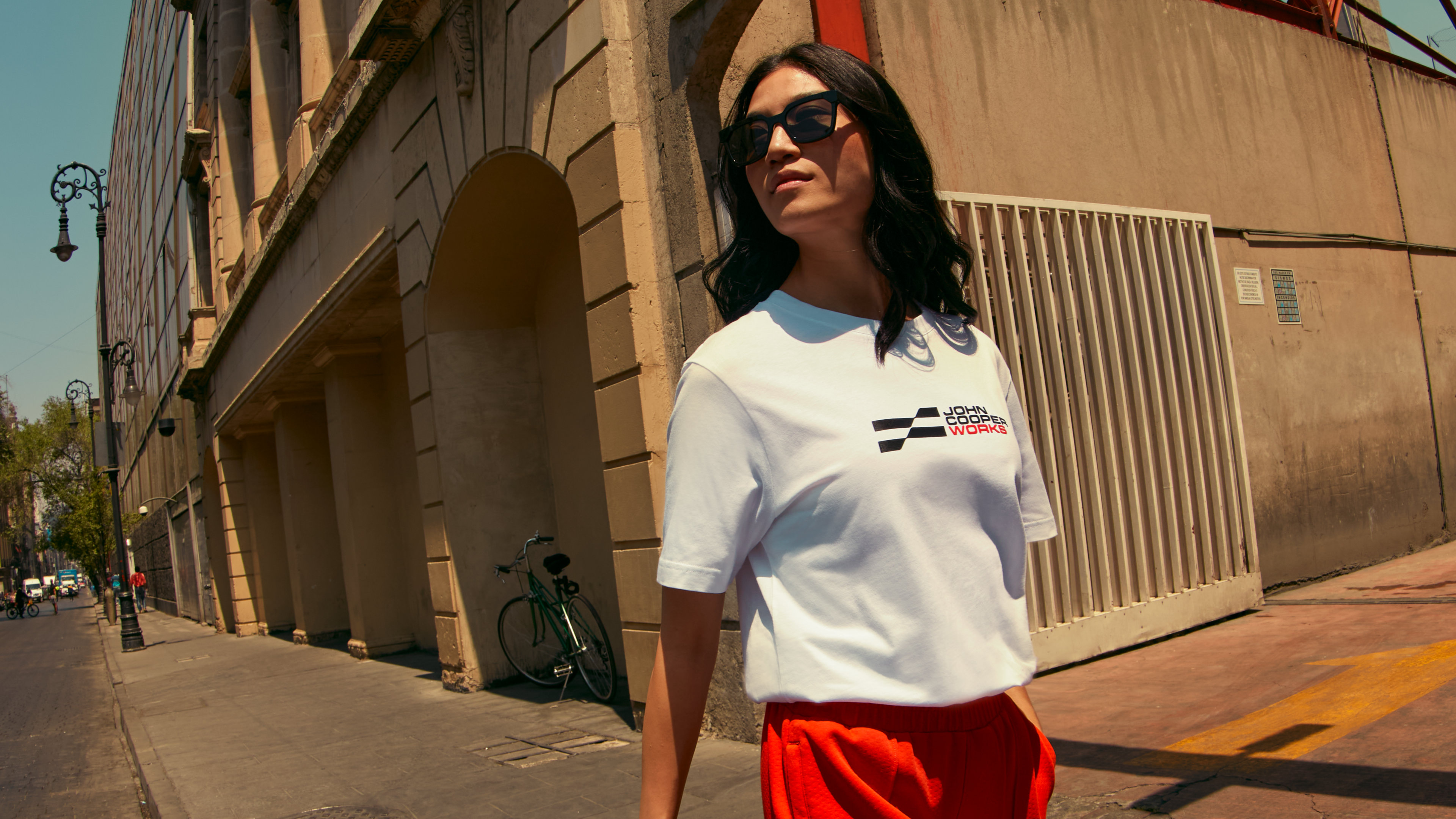 A woman in sunglasses and a John Cooper Works t-shirt walks beside a brown stone building