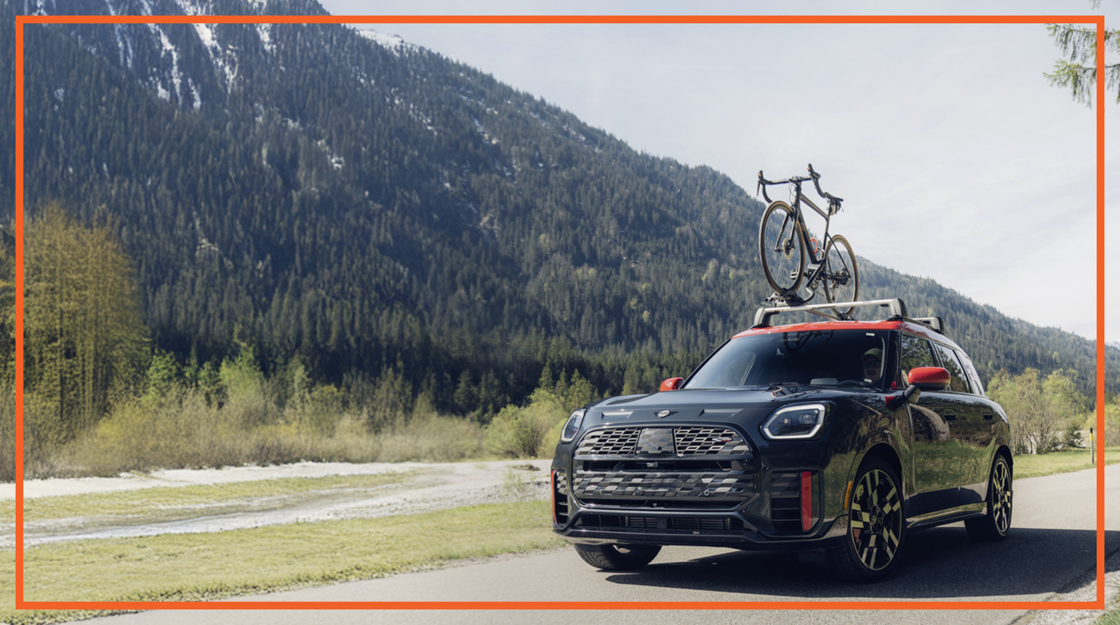 A MINI JCW equipped with a roof rack carrying a bicycle drives along a mountainside road.