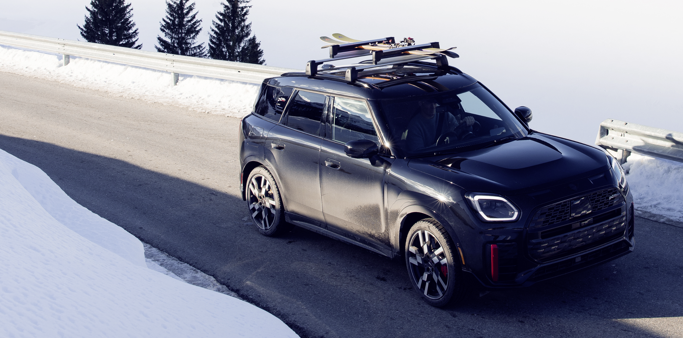 A black MINI Countryman with skis mounted on the roof, drives along a mountain road. White snow blankets the ground on both sides of the road and up the tree covered mountain in the background.